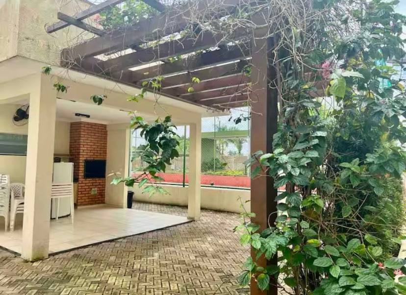 An outdoor seating area is framed by a wooden pergola adorned with climbing plants. A smooth tiled floor extends beneath, with a small television mounted on a brick wall. Nearby, a tennis court is visible through the foliage, enhancing the tranquil ambiance.