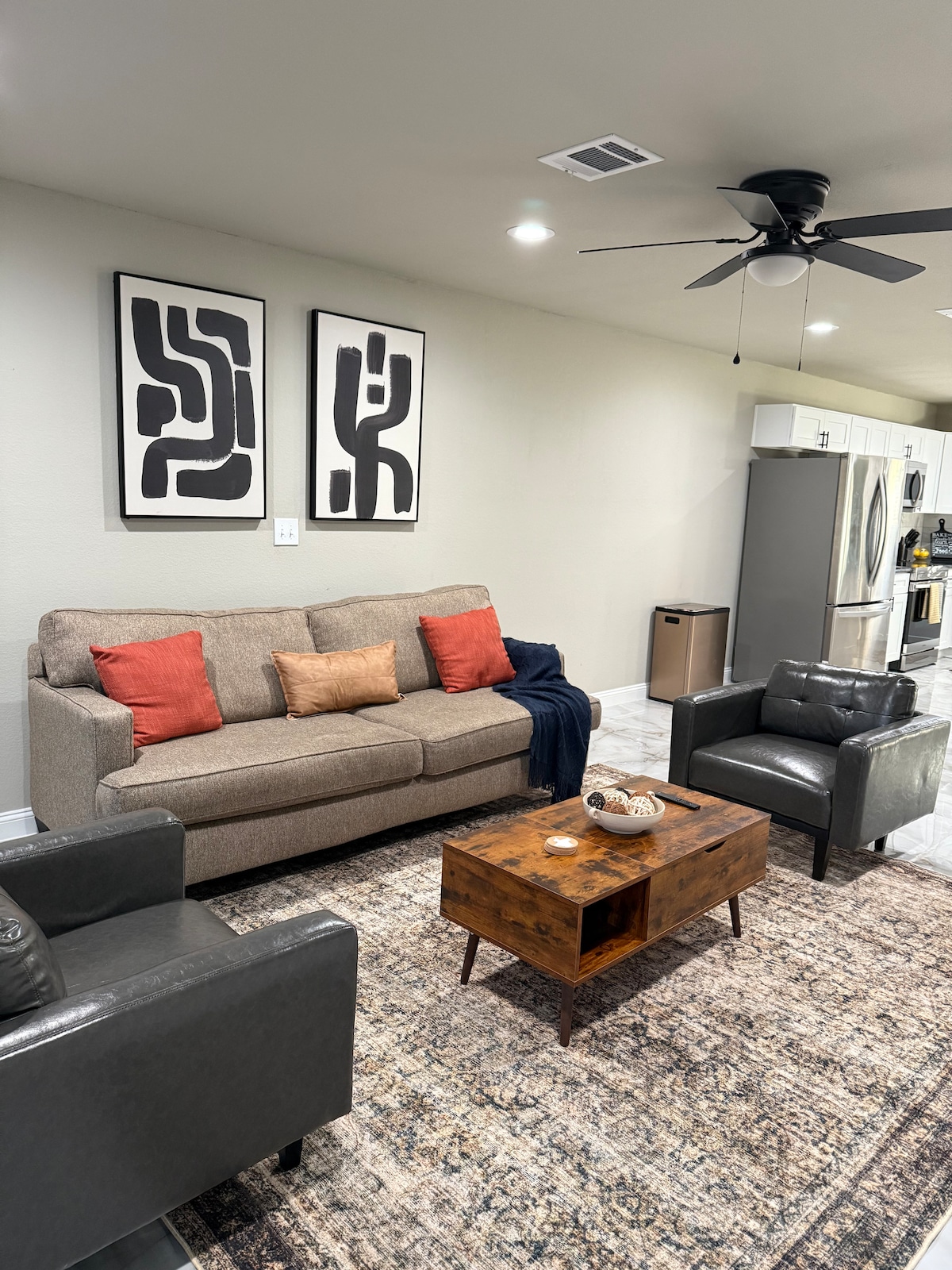 A welcoming living area is presented with a sofa adorned with colorful cushions, accompanied by two darker armchairs. A wooden coffee table sits center, and abstract art pieces are displayed on the wall above the sofa, enhancing the modern decor and open concept layout.