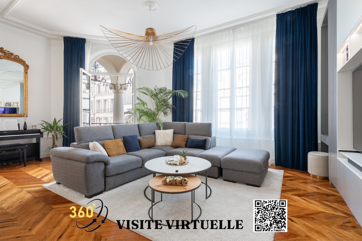 A spacious living area features a large sectional sofa adorned with cushions, positioned near a round coffee table. Tall windows are draped with deep blue curtains, allowing ample natural light. A light fixture with a unique design adds interest above, and a piano is positioned in one corner.
