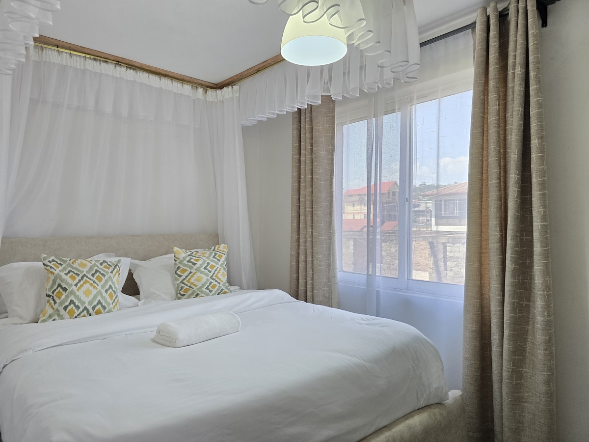 A cozy sleeping area features a spacious bed draped with light, sheer fabric. Two decorative pillows with geometric patterns add color. Sunlight streams through large windows, where delicate curtains softly frame the view outside.