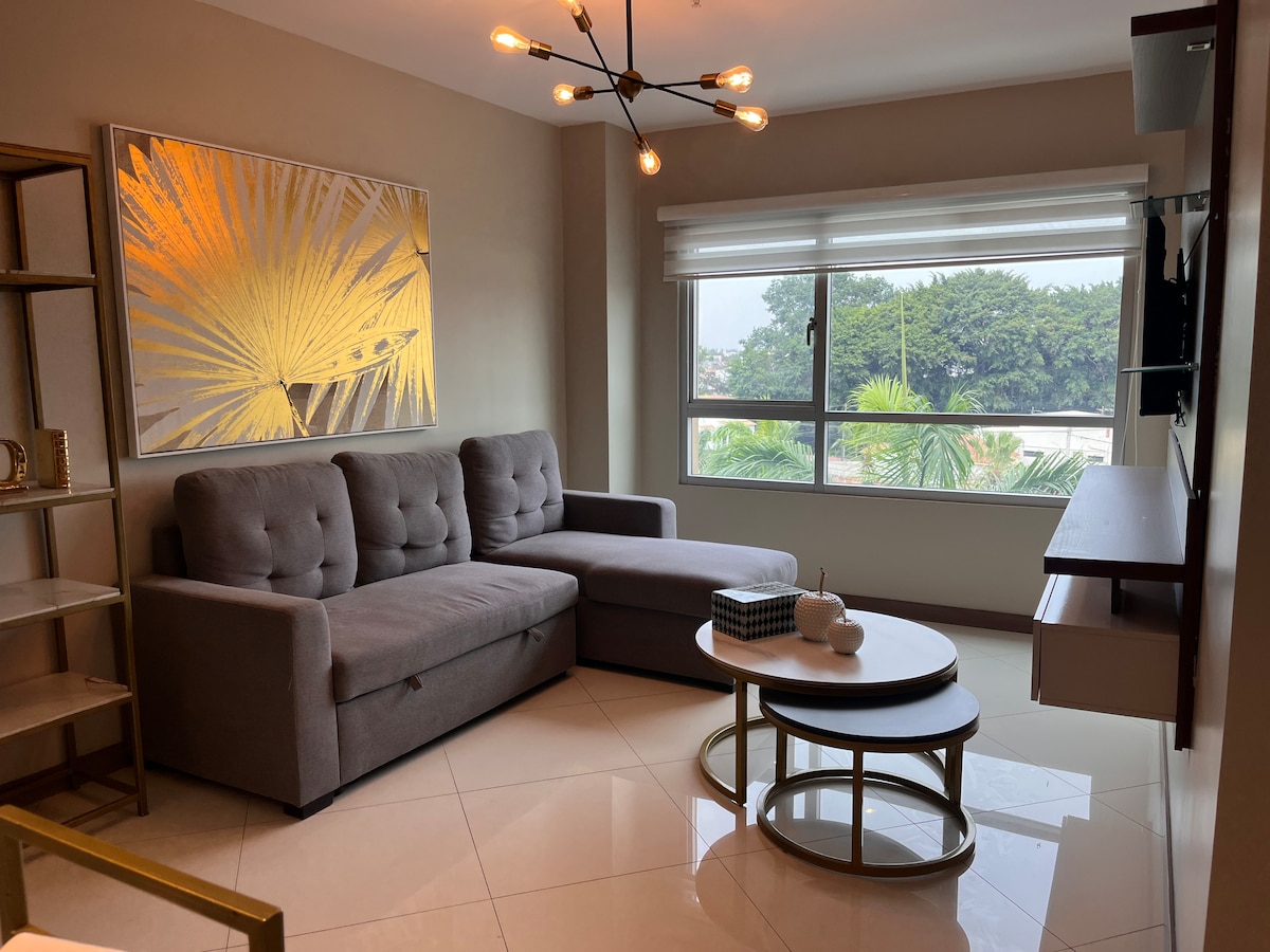 A modern living area features a gray sectional sofa positioned beside a decorative wall with an elegant gold art piece. Natural light floods through a large window, illuminating the polished tile floor and a round coffee table placed in front.