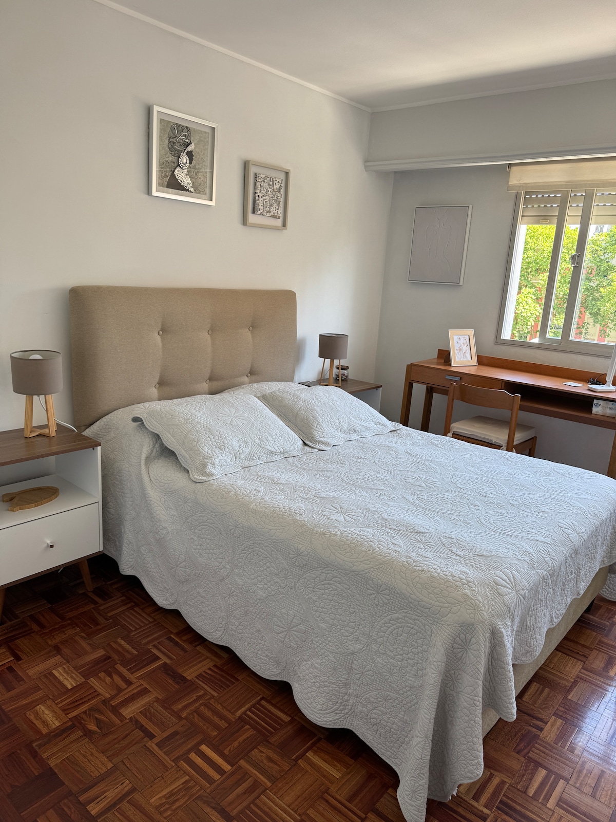 A bright bedroom features a spacious bed dressed in a light blue quilt. Two bedside tables with lamps and a wooden work desk are positioned near a window, allowing natural light to illuminate the room. Wall art adds a personal touch to the serene environment.