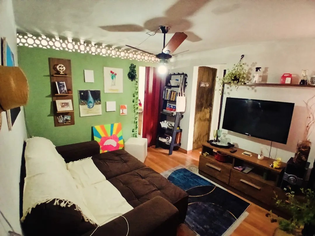 A cozy living area features a brown sectional sofa draped with a light throw. Green accent walls are decorated with various artworks arranged on shelves. Natural light enters from a nearby window, with a ceiling fan providing ventilation. A media console holds a television and decorative items.