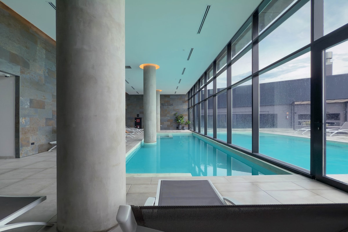 An indoor swimming pool is featured, surrounded by large glass windows that allow natural light to fill the space. A light-colored tile floor complements the pool's clear, blue water. Comfortable lounge chairs line the poolside, and a concrete pillar offers structural support.