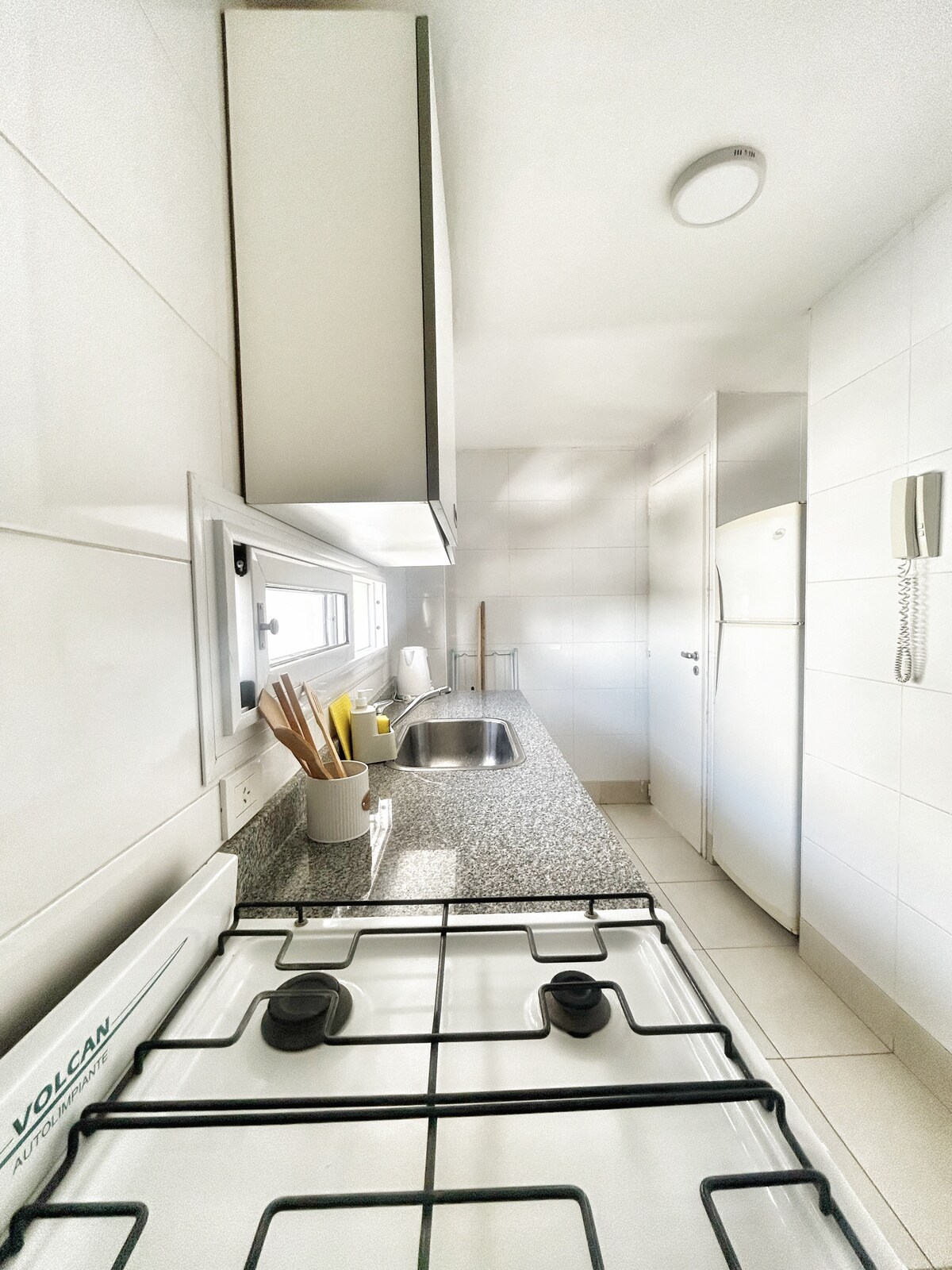 A modern kitchen is presented with sleek white cabinetry and a granite countertop. A gas stove with two burners is visible, alongside a double-basin sink. Natural light filters through a nearby window, illuminating the space. A refrigerator is positioned against the wall, adding functionality.