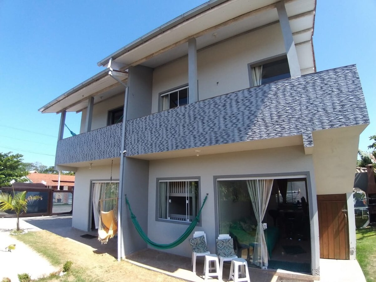 A two-story house features a textured exterior with a shaded balcony above and large windows below. A hammock hangs on the porch, accompanied by two white chairs. A grassy area and pathway lead to the entry door, framed by surrounding greenery.