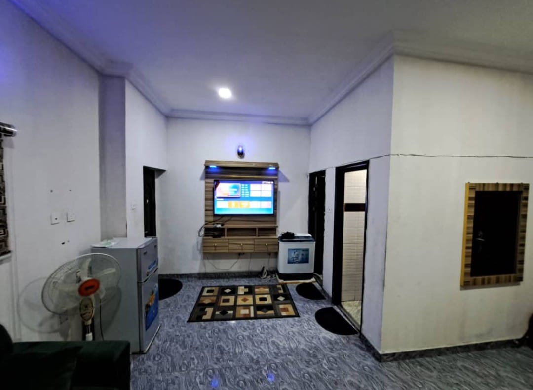 A living area is featured with a flat-screen television mounted on a wall. A patterned rug covers the floor, while comfortable seating is provided in a cozy arrangement. A small refrigerator and a fan are also visible, contributing to a functional and inviting space.
