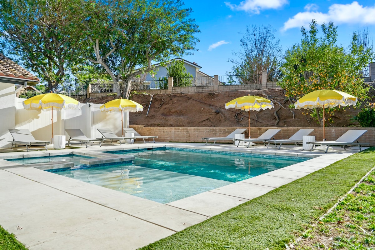 The backyard features a private pool with steps leading into the water. Sun loungers are arranged around the pool, accompanied by yellow umbrellas providing shade. Lush green grass borders the area, and surrounding landscaping adds a natural touch.