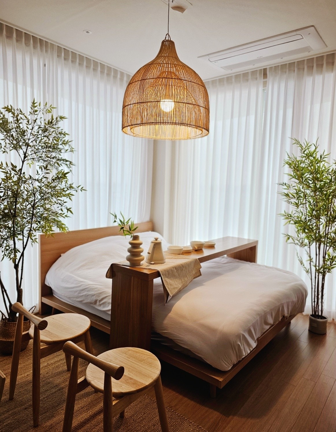 A cozy bedroom features a spacious bed dressed in soft white linens, complemented by natural wood furnishings. Light filters through sheer white curtains, enhancing the calming atmosphere. Two wooden stools are positioned near a small decorative table, and tall green plants add a touch of nature.
