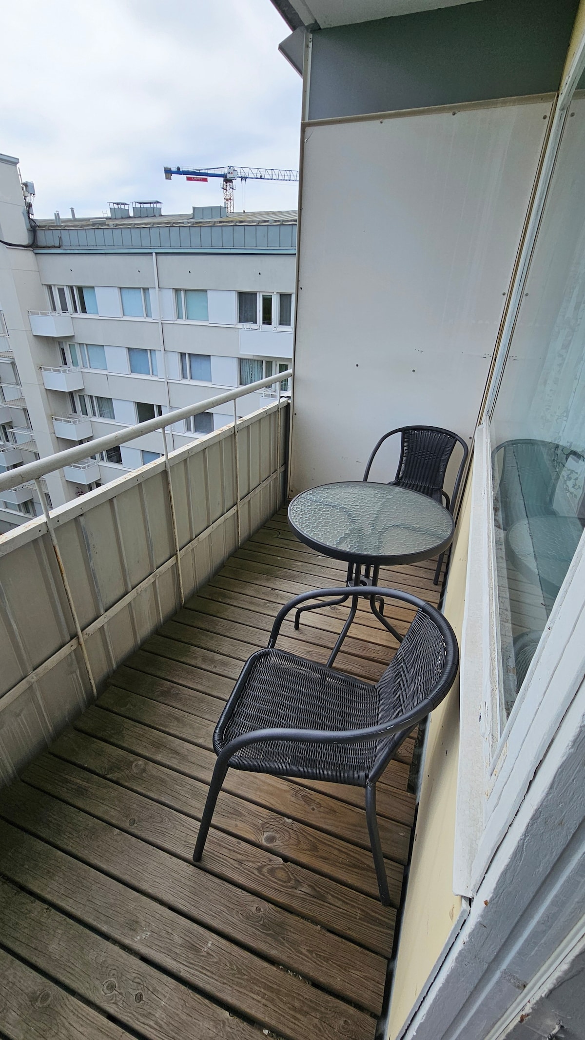 A small balcony features wooden flooring and a glass-top table with two black chairs. The surrounding glass railing provides an unobstructed view. Nearby buildings are visible in the background, with construction equipment seen in the distance.