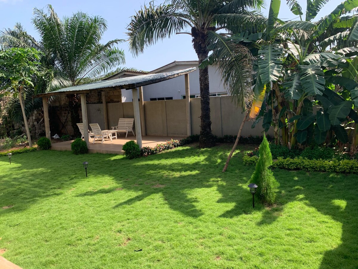 A lush green garden features neatly trimmed grass and various tropical plants. Two lounge chairs are situated under a shaded area, providing a perfect spot for relaxation. Soft outdoor lighting is visible, enhancing the calm ambiance of the space.