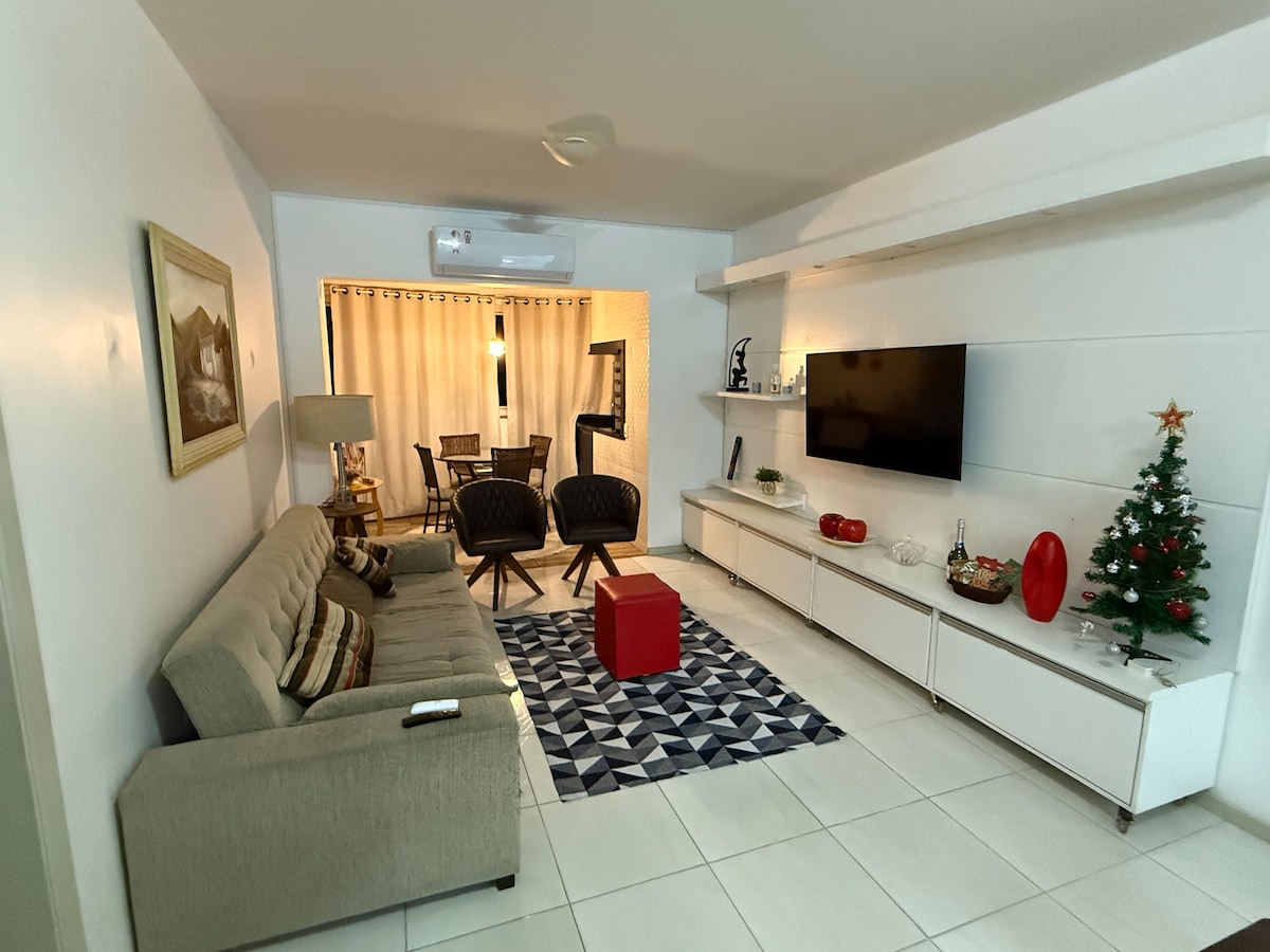 A modern living room features a comfortable sofa and accent chairs arranged around a geometric patterned rug. A flat-screen TV is mounted on the wall, and a small red ottoman adds a pop of color. Natural light filters through sheer curtains, creating a welcoming atmosphere.