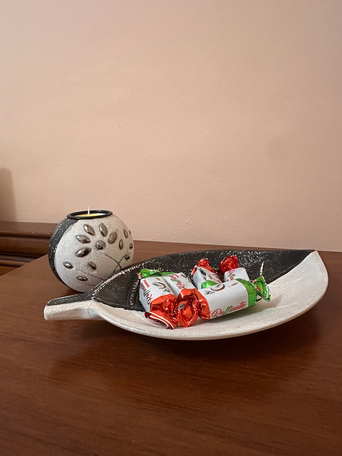 A decorative bowl is displayed on a wooden surface, holding an assortment of wrapped candies in bright colors. The bowl features a unique leaf shape, with a contrasting black and white design, enhancing the simple elegance of the setting.