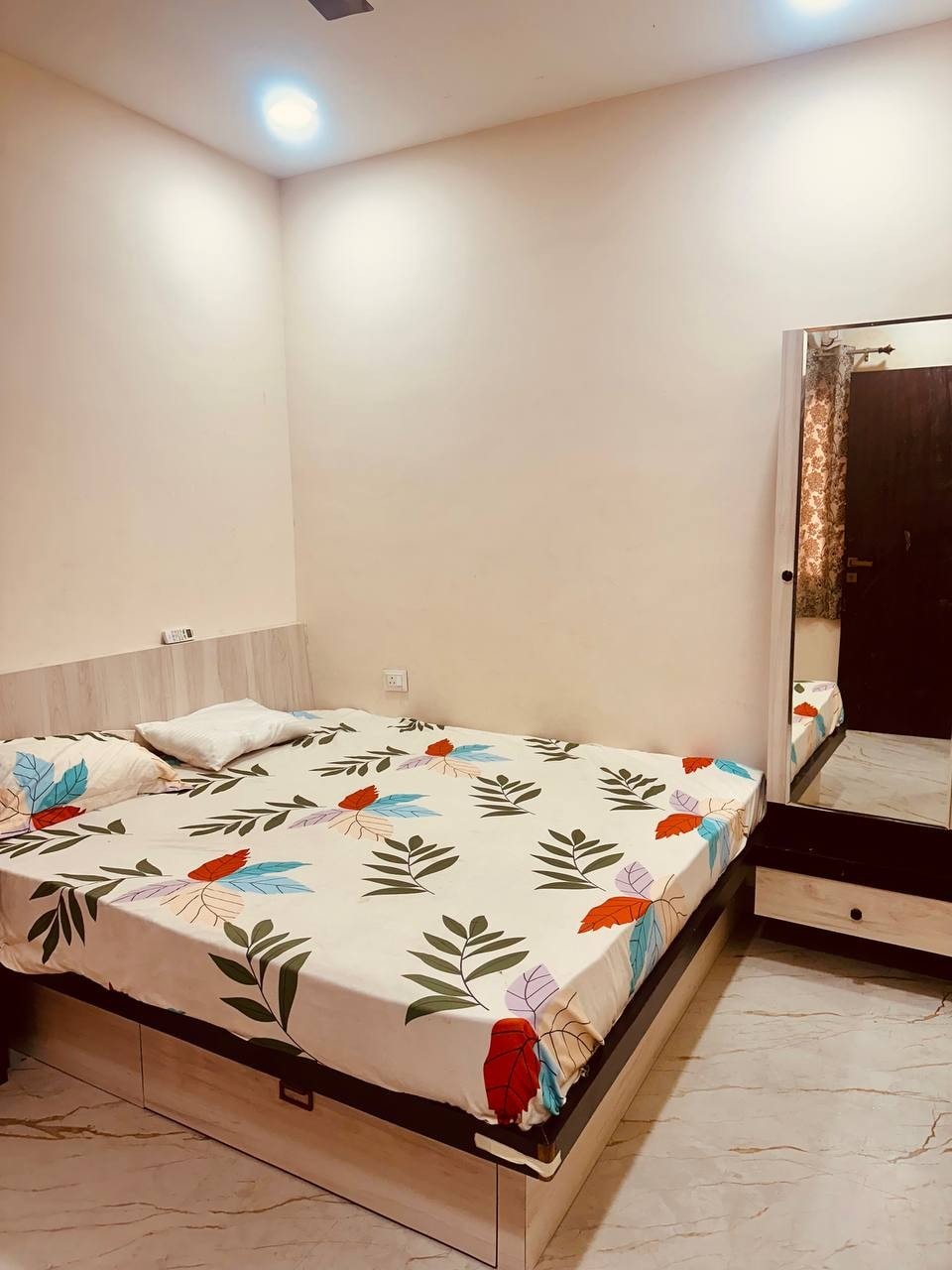 A cozy sleeping area features a spacious bed adorned with a patterned bedspread showcasing leaves and flowers. A mirror reflects the room's light hues. Natural light filters through curtains, creating a calming atmosphere. Additional furnishings include a bedside table and storage below the bed.