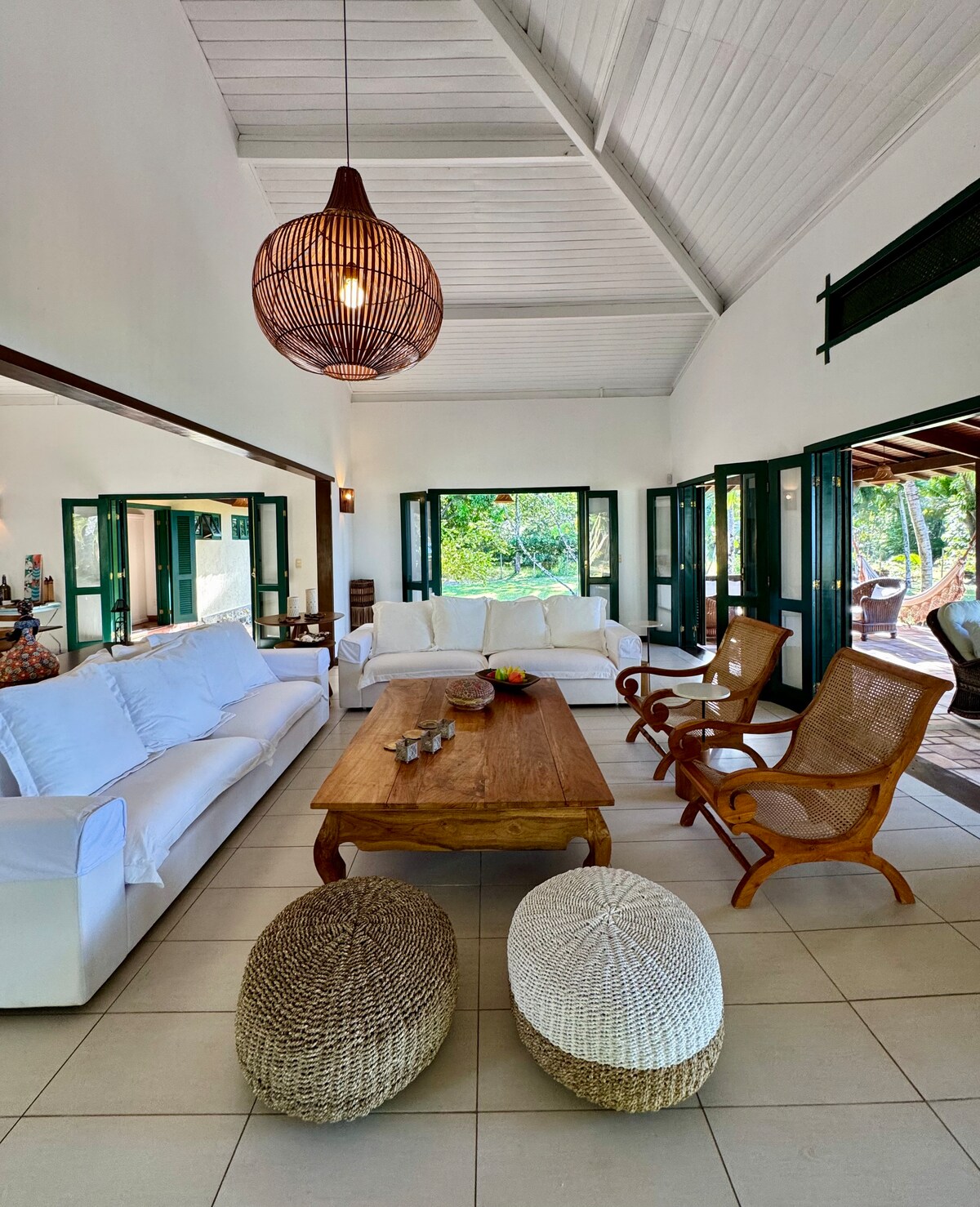A bright living area features large windows that open to the outside, allowing natural light to fill the space. Comfortable white sofas are arranged around a wooden coffee table, with woven poufs nearby. A statement light fixture hangs from the high ceiling, adding warmth to the setting.