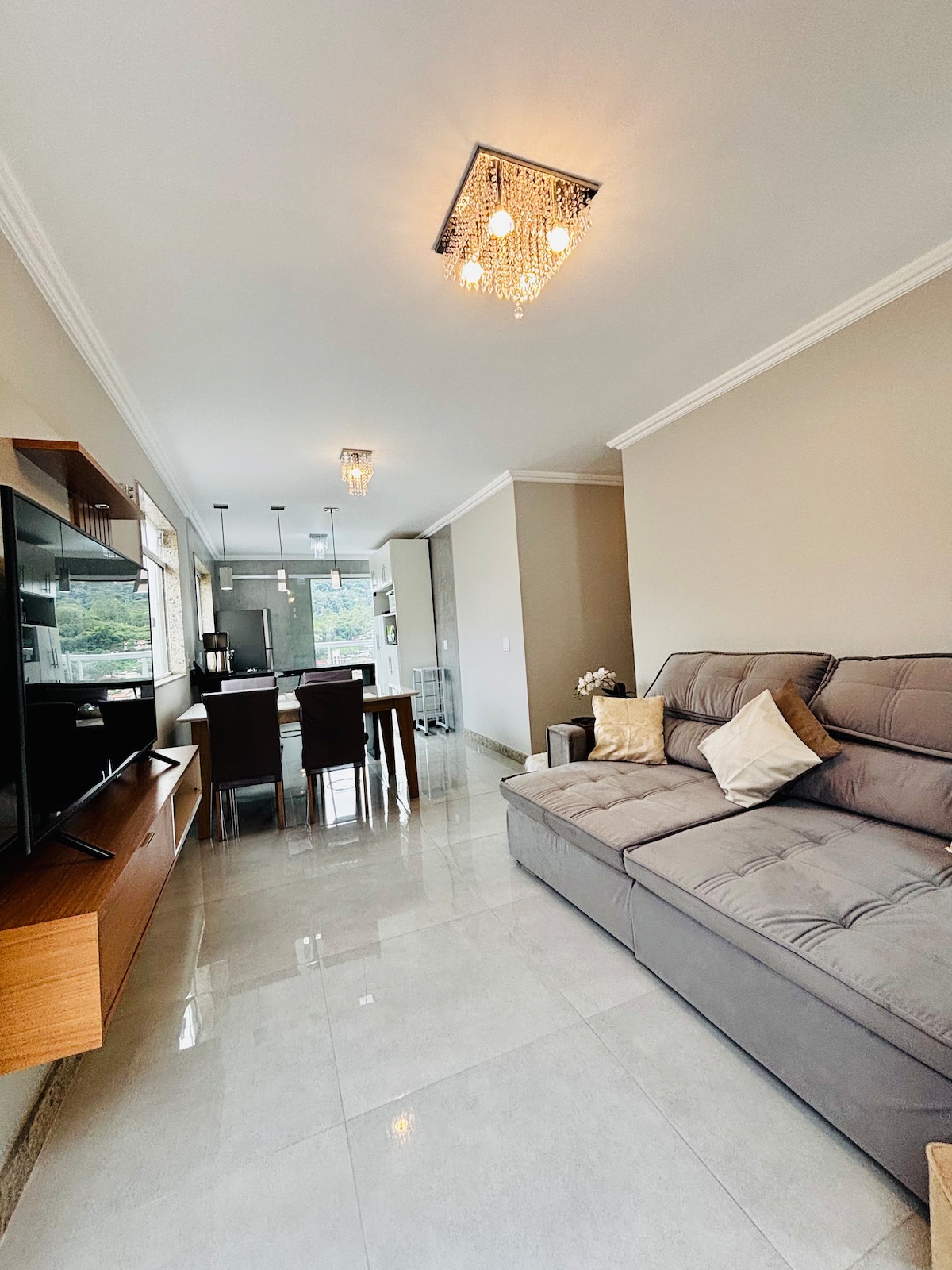 A modern living area features a plush gray sofa with decorative pillows, alongside a wooden entertainment unit. A dining table with chairs is visible in the background, complemented by overhead lighting and a view of the outdoors through large windows.