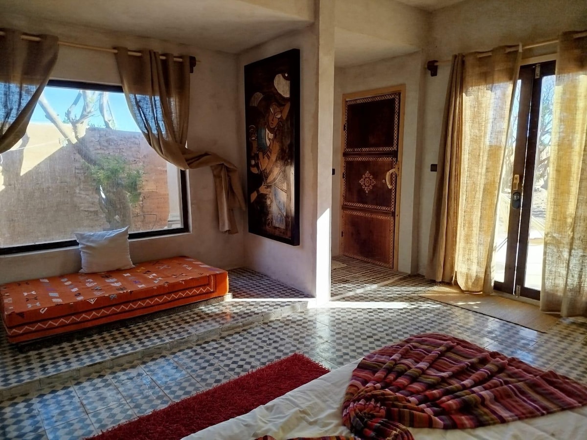 A sunlit room features a cozy sleeping area with a patterned blanket draped over a spacious bed. A vibrant orange seating area is positioned by a large window adorned with light curtains. Artistic wall decor and natural light enhance the room's ambiance.