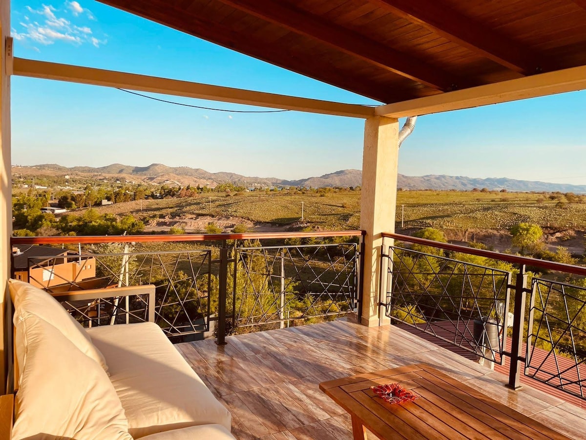 A spacious outdoor terrace features comfortable seating and a wooden table, overlooking sprawling green landscapes and mountainous scenery. The setting sun casts a warm glow, enhancing the relaxed vibe of the area. The terrace is surrounded by a secure railing, providing safety and unobstructed views.