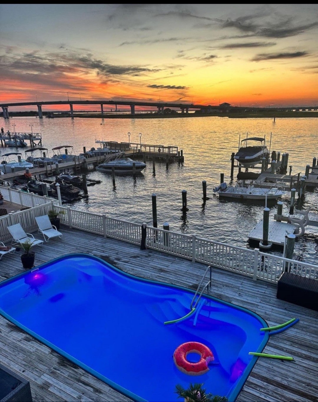 A private pool sits on a deck, overlooking a vibrant bay at sunset. The pool features a colorful float and is surrounded by lounge chairs. In the distance, boats are docked at the marina, with a bridge visible against the colorful evening sky.