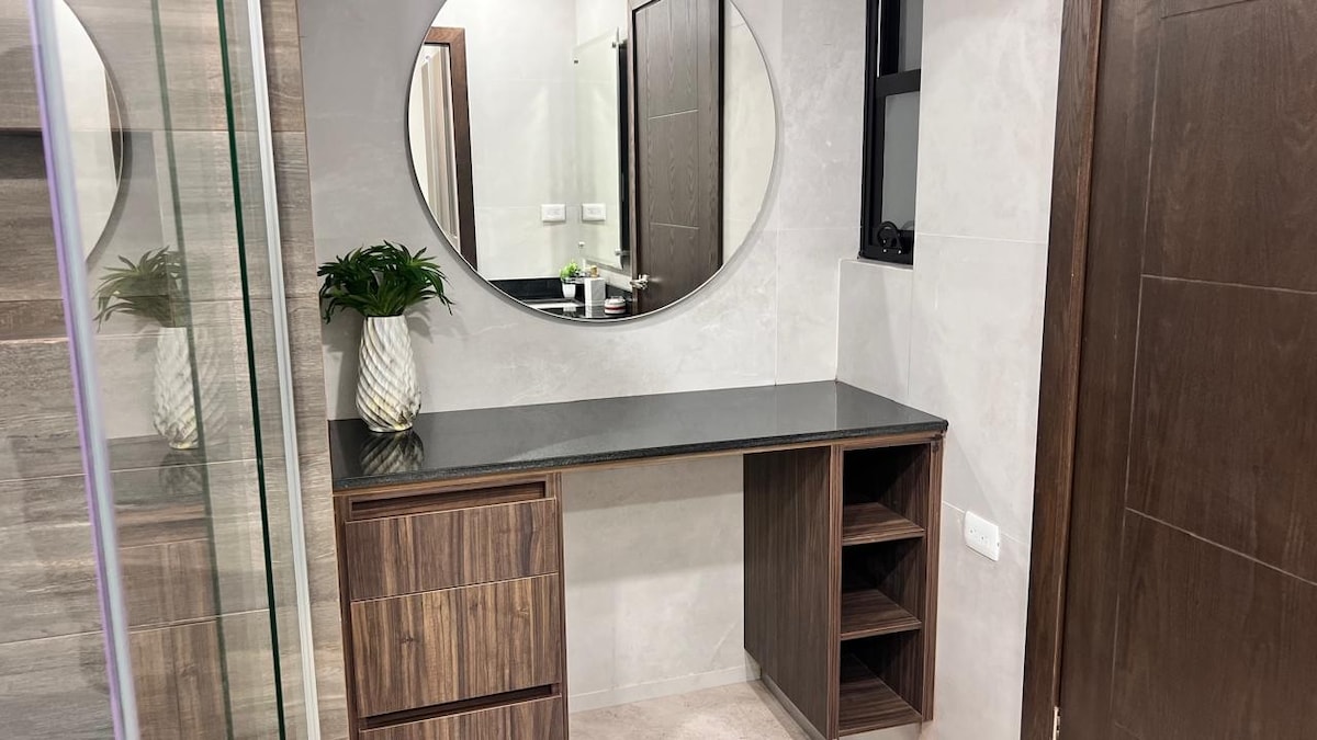 A modern bathroom space is portrayed, featuring a circular mirror above a dark stone countertop. Wooden cabinetry provides storage, including a set of drawers and open shelves. The wall is finished with light-colored tiles, enhancing the sleek design.