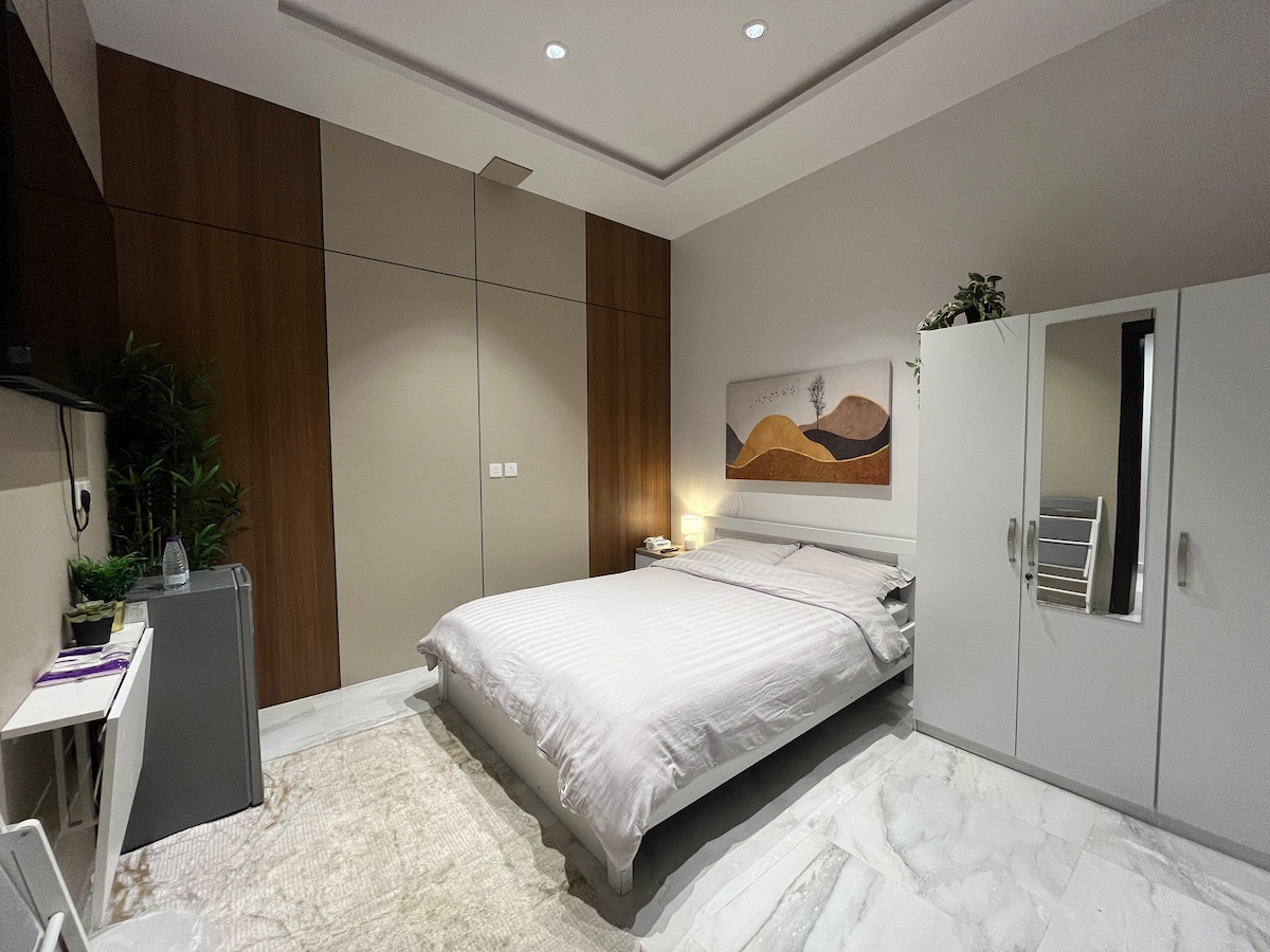 A spacious bedroom features a queen-size bed with light-colored bedding and a simple wall art above. A wardrobe with sliding doors and a small desk are visible, while soft lighting from a bedside lamp creates a calming ambience. The room’s floor is finished with light marble.
