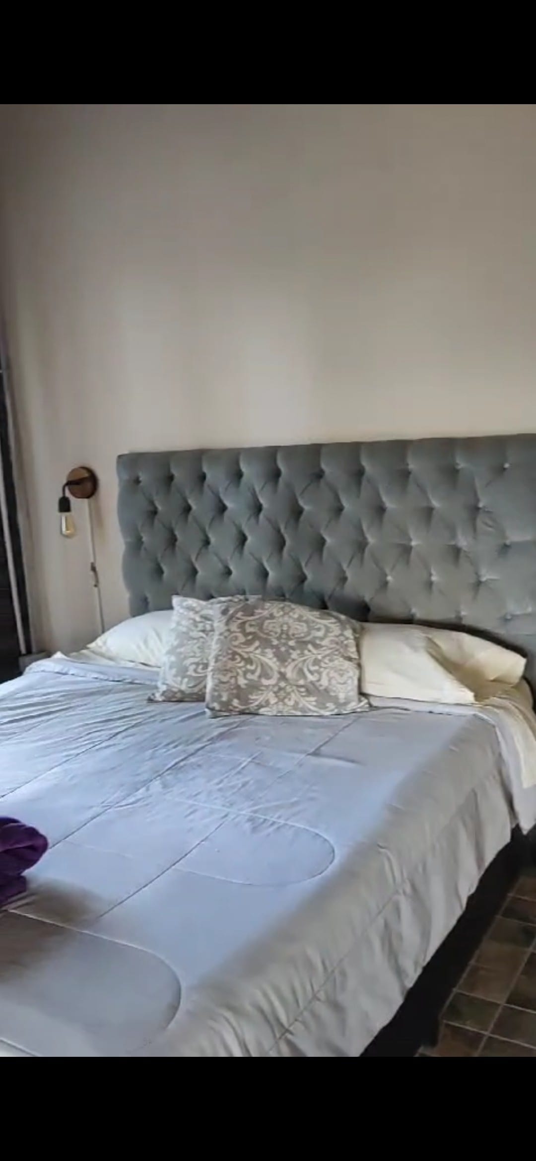 A spacious sleeping area features a plush, tufted headboard in a soft grey tone. Decorative pillows add texture to the bedding, which is neatly arranged. Natural light filters through large windows, illuminating the room.