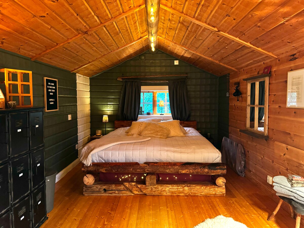 A cozy sleeping area features a spacious bed with a light-colored comforter, set against wooden walls and a high ceiling. Soft lighting from a bedside lamp casts a warm glow, while dark curtains frame a window that allows natural light to enter.