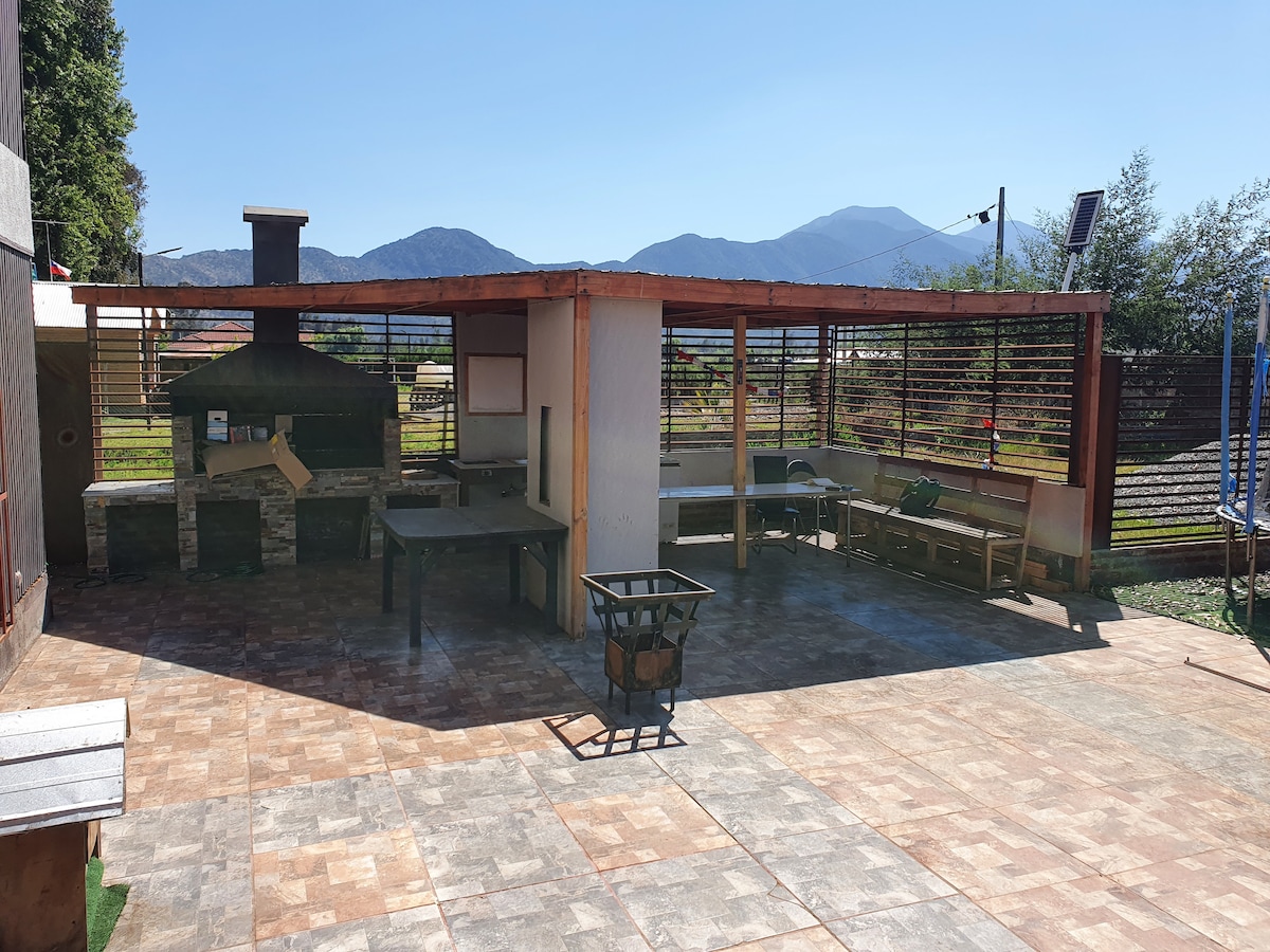 An outdoor grilling area features a built-in stone oven and a spacious countertop. Several benches are arranged around a large patio, with mountains visible in the background. Natural light creates a bright and open atmosphere.