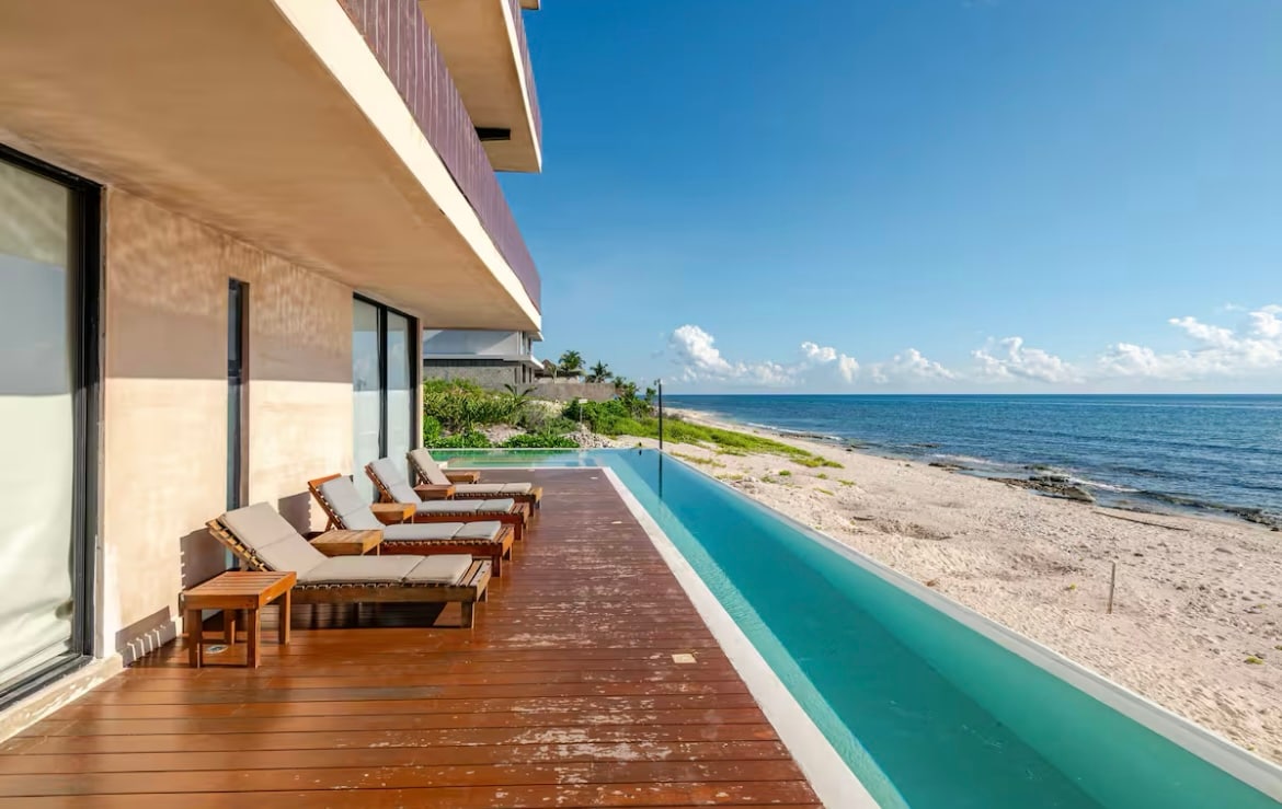 A wooden deck is presented with several lounge chairs, overlooking a tranquil infinity pool that seamlessly meets the sandy beach and ocean beyond. The scene is complemented by a clear blue sky, enhancing the coastal ambiance of the space.