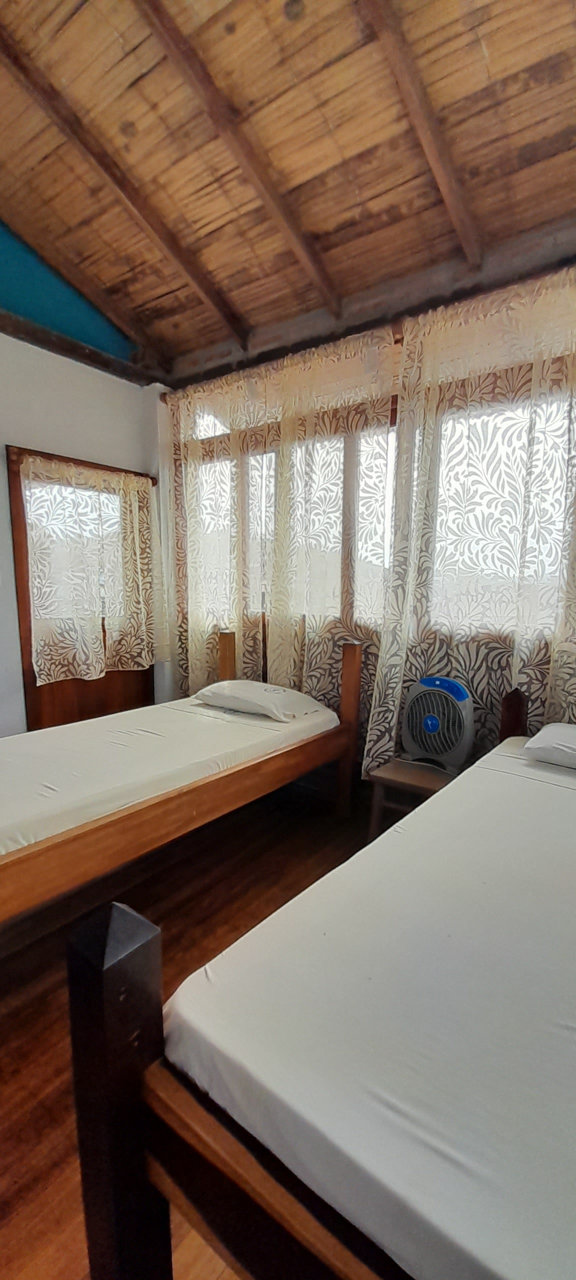 A room is presented with two simple beds featuring light bedding and wooden frames. Large windows are adorned with lace curtains, allowing natural light to fill the space. A portable fan is placed beside one bed, and warm wooden floors are visible throughout.