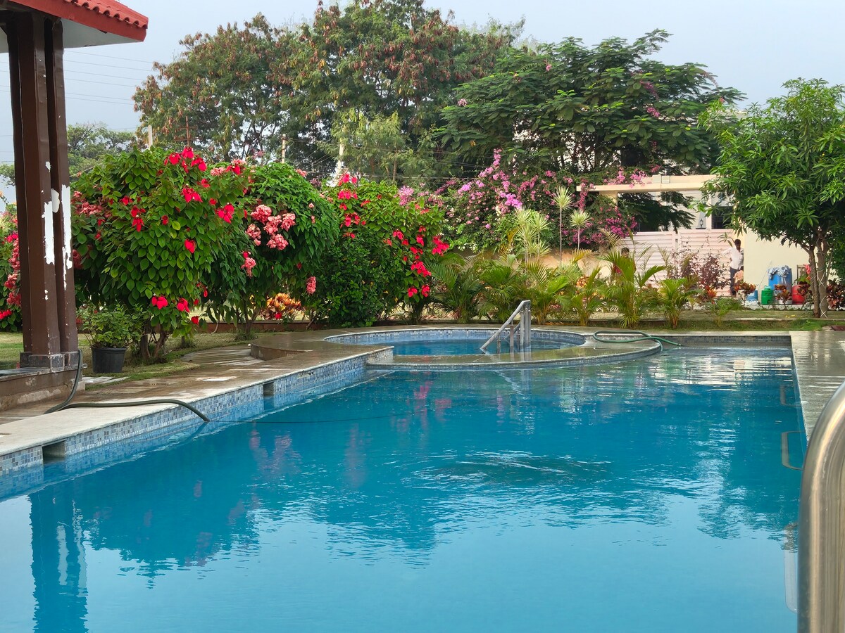 A clear swimming pool reflects the surrounding vibrant greenery and flowering plants. The pool area is complemented by a small outdoor structure, and spaces for relaxation are visible along the edge, inviting a serene atmosphere to the setting.