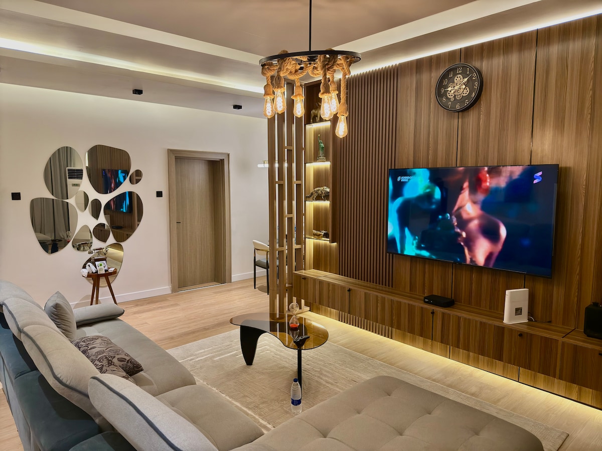 A contemporary living room is featured with a comfortable sectional sofa and a stylish coffee table. A large wall-mounted television is positioned in front, with decorative wooden paneling and ambient lighting enhancing the atmosphere. A circular mirror reflects the space, adding visual interest.