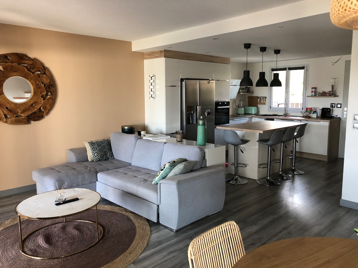 An open-plan living space is displayed, featuring a comfortable gray sectional sofa with accent cushions. A round coffee table sits on a textured area rug. The modern kitchen is equipped with sleek appliances and bar stools, offering a sense of casual dining and entertaining.