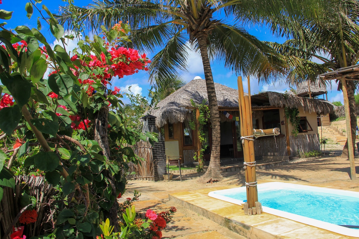 A cozy bungalow is nestled among palm trees, showcasing thatched roofing and a tranquil ambiance. A small swimming pool is positioned nearby, surrounded by vibrant flowers and greenery, creating a serene outdoor space ideal for relaxation.