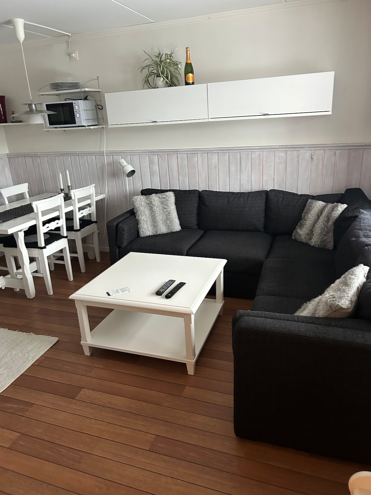 A living area is presented with a dark sectional sofa featuring two decorative cushions. A white coffee table occupies the center, and a dining table with four chairs is visible in the background. A microwave and other kitchen items are located on the wall shelf, creating a functional space.