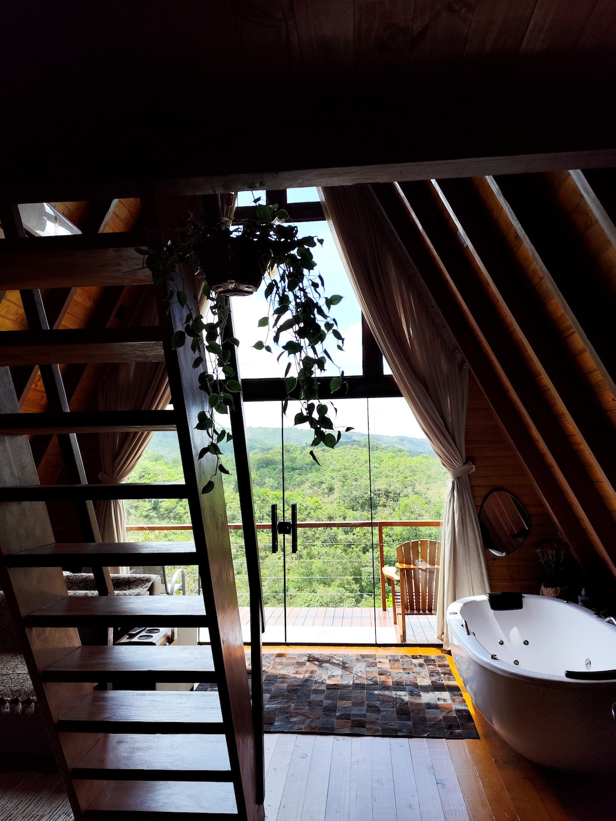 A staircase leads to a cozy loft area with wooden beams and large windows, offering views of the surrounding greenery. A freestanding bathtub is positioned near the windowsill, accompanied by a small chair on the deck outside. Decorative plants add a natural touch to the space.