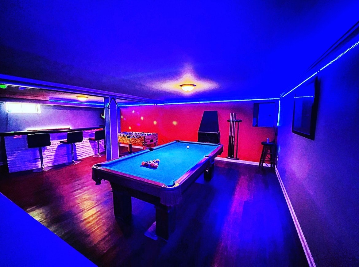 A vibrant basement game area is illuminated by soft blue lighting. A pool table is positioned at the center, surrounded by stylish seating at a bar counter in the background. The walls are adorned with cues and there is a cozy ambiance throughout the space.