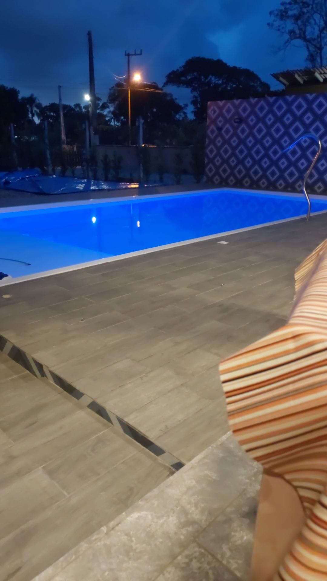 A serene pool area is highlighted by smooth, tiled surfaces that surround the water. The pool is illuminated with soft blue lighting, creating a calming effect as the evening sets in. A nearby lounge chair features a striped pattern, enhancing the inviting space.