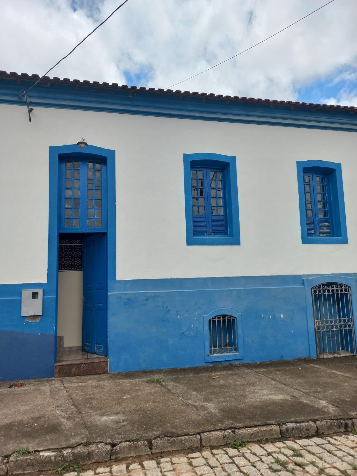 A charming building with a white exterior is accented by blue details, including window frames and a door. The entrance features a decorative gate, and the ground is paved with stone. The clouds above add a dynamic element to the scene.