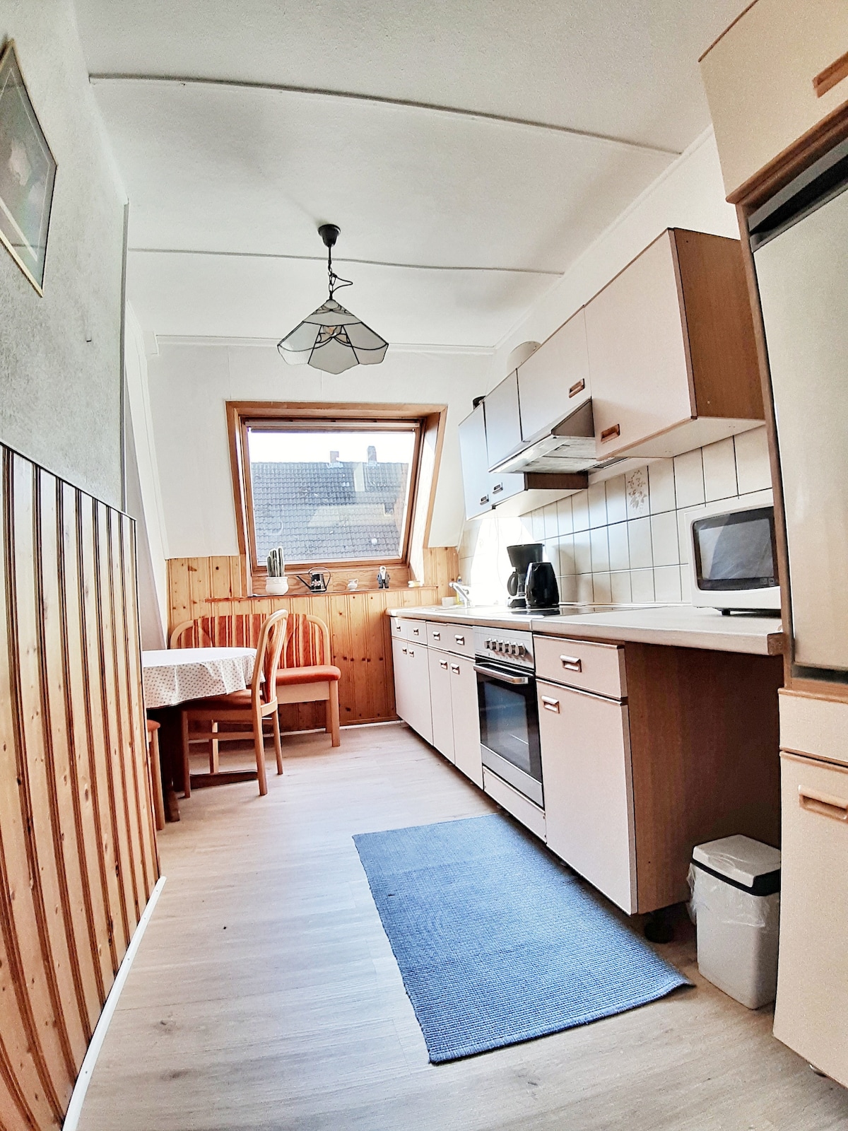 A bright kitchen is featured, showcasing wood paneling and modern cabinetry. A dining table with two wooden chairs sits beside a large window, allowing natural light to fill the space. Equipped appliances, including a stove and microwave, are visible, along with a small rug on the floor.