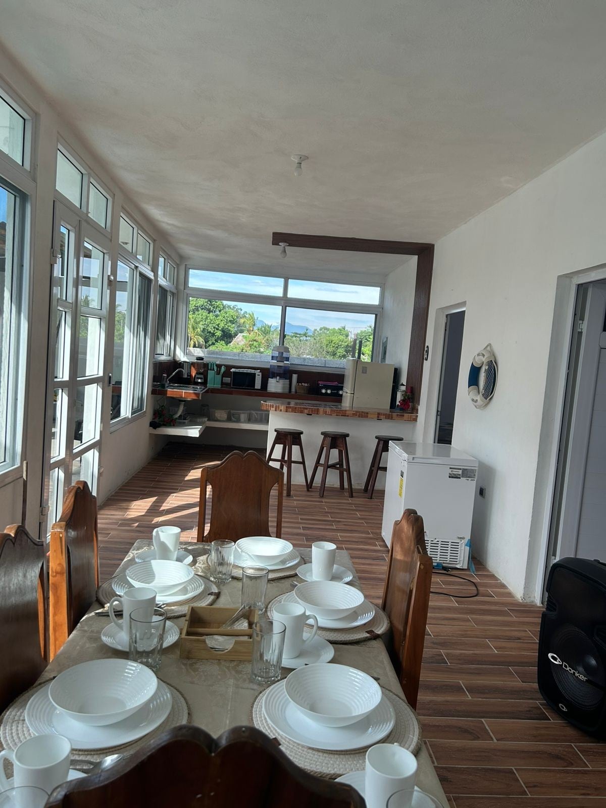 A dining area features a wooden table set for a meal, complemented by white plates and clear glassware. Large windows surround the space, allowing natural light to illuminate the room. A kitchen area is visible in the background, equipped with kitchen appliances.