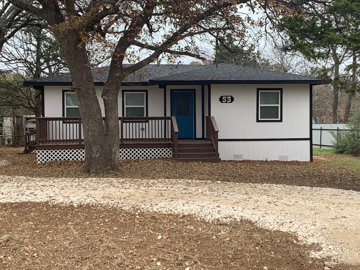 A single-story house with a dark blue front door is visible, surrounded by trees. The exterior is painted white with dark trim. A gravel driveway leads to the entrance, which features a small covered porch with wooden railings.