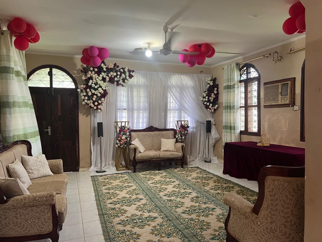 The living area is spacious and features a floral arrangement at the entrance, complemented by decorative balloons. A plush seating arrangement includes a sofa and armchairs, arranged around an area rug. Natural light fills the room through large windows adorned with sheer curtains.
