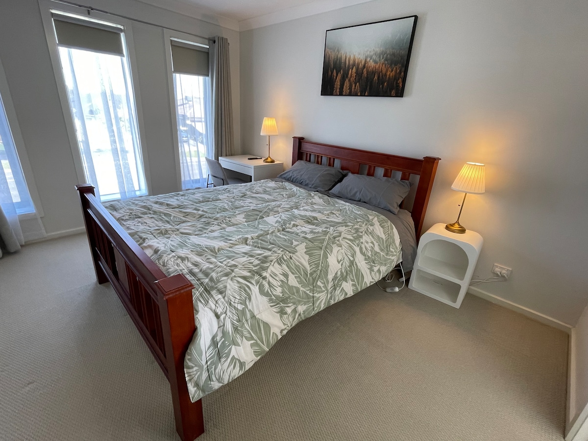 A cozy bedroom features a wooden bed frame with a patterned comforter and two accent pillows. Nightstands with lamps are placed on either side of the bed. Natural light streams through large windows, highlighting a serene atmosphere.
