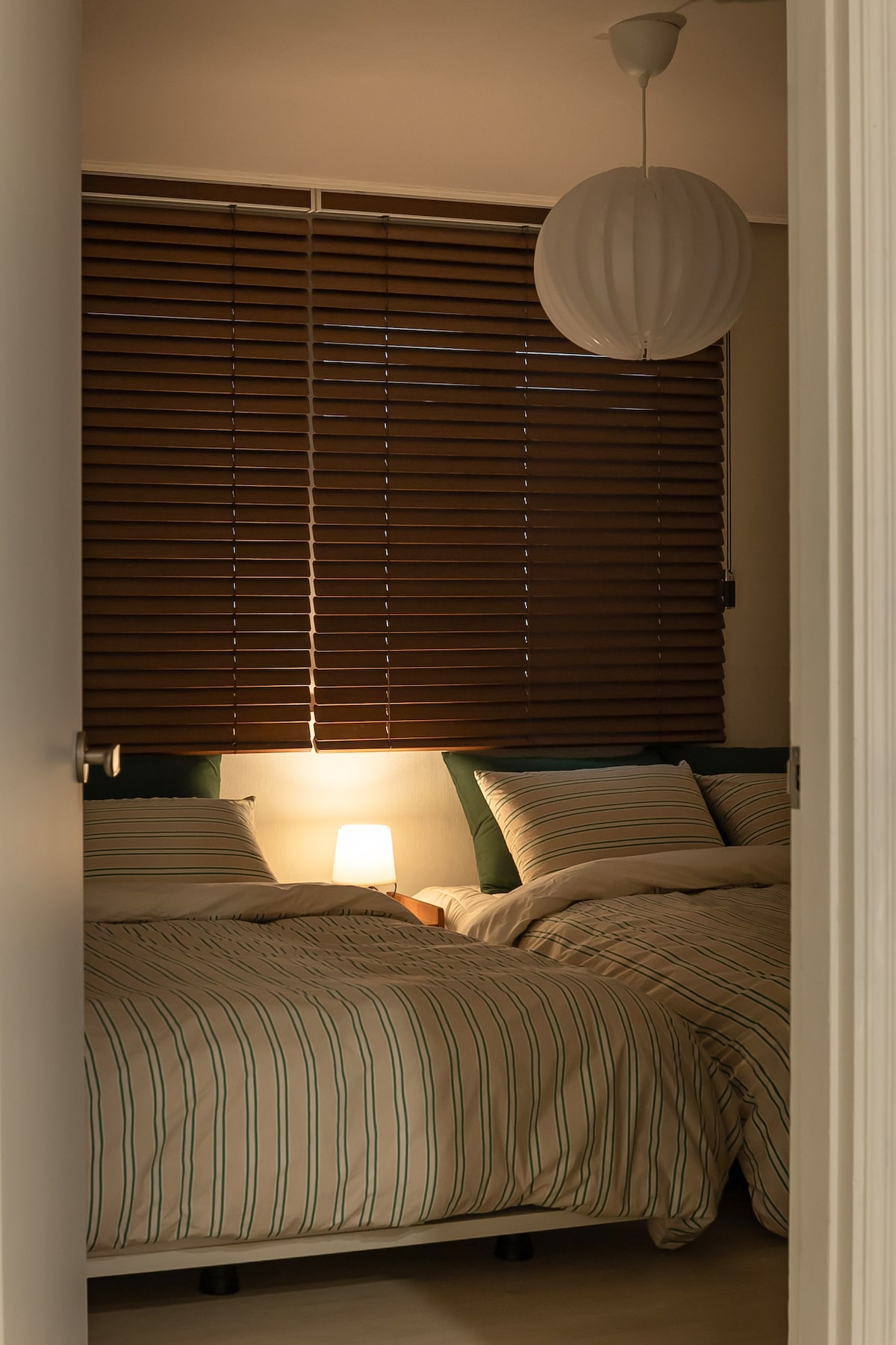 A cozy bedroom is depicted, featuring two spacious beds dressed in soft striped linens. A warm bedside lamp provides gentle illumination, and the window is adorned with wooden blinds, softening the ambient light in the room.