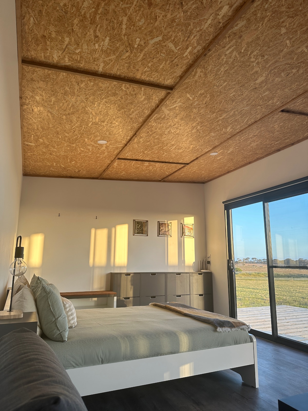 A modern bedroom features a spacious bed with neutral bedding. Large sliding doors allow natural light to enter, revealing views of the landscape. An accent chair and a dresser are present, while framed artworks adorn the walls, contributing a subtle touch of decor.