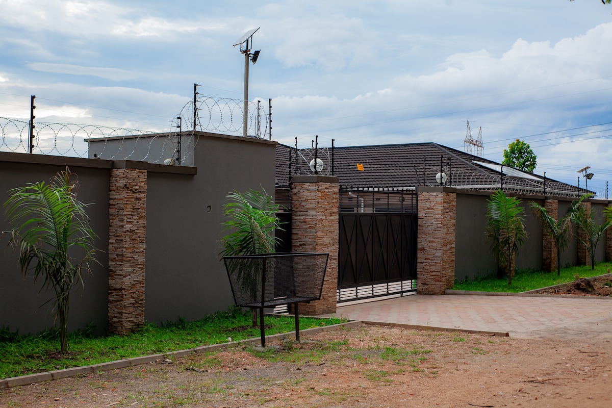 A secure entrance is framed by a tall stone wall with barbed wire atop. A solid black gate stands at the center, surrounded by well-maintained grass and young palm trees, contributing to a sense of privacy and safety.
