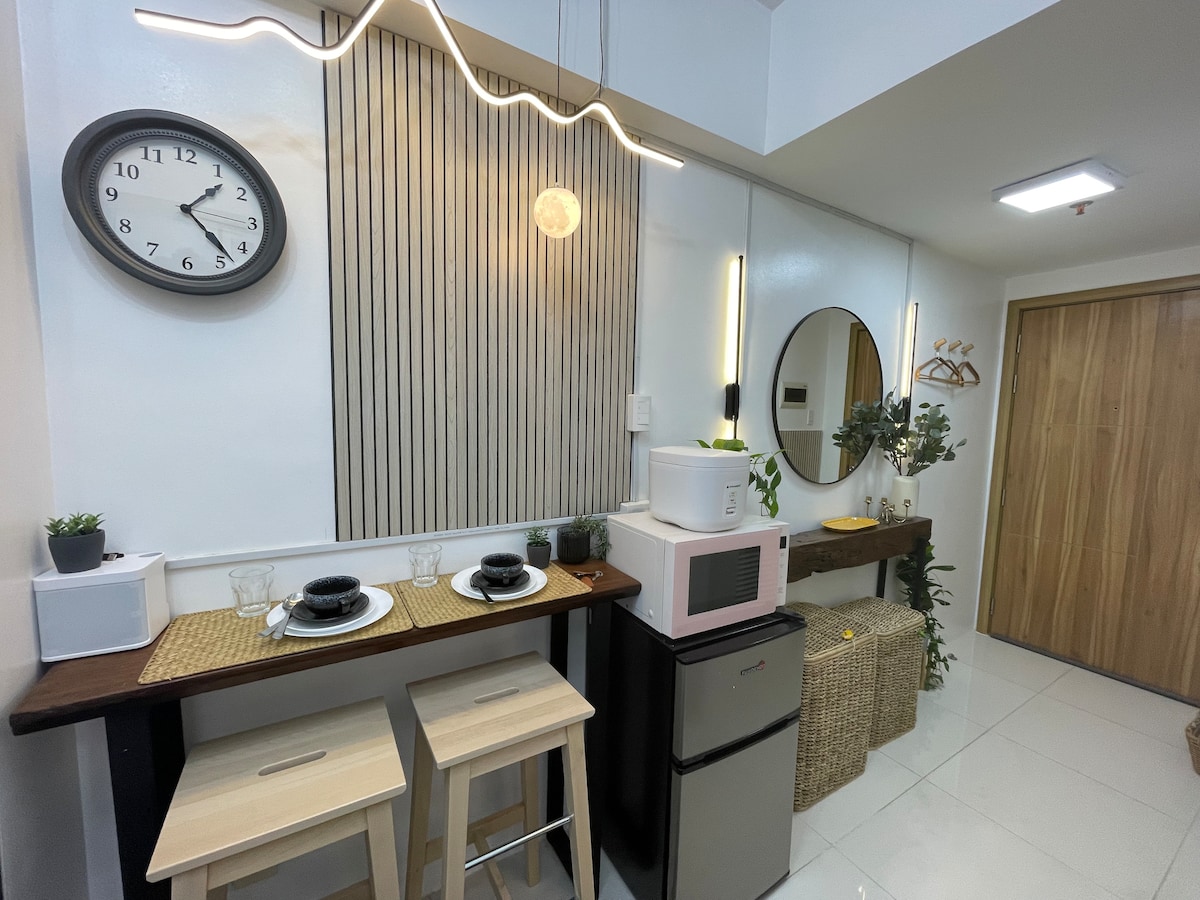 A compact kitchenette area features a wooden bar table with two stools, accompanied by two place settings. A microwave and refrigerator are visible, while a mirror and decorative items enhance the space. Soft lighting creates a welcoming ambiance.