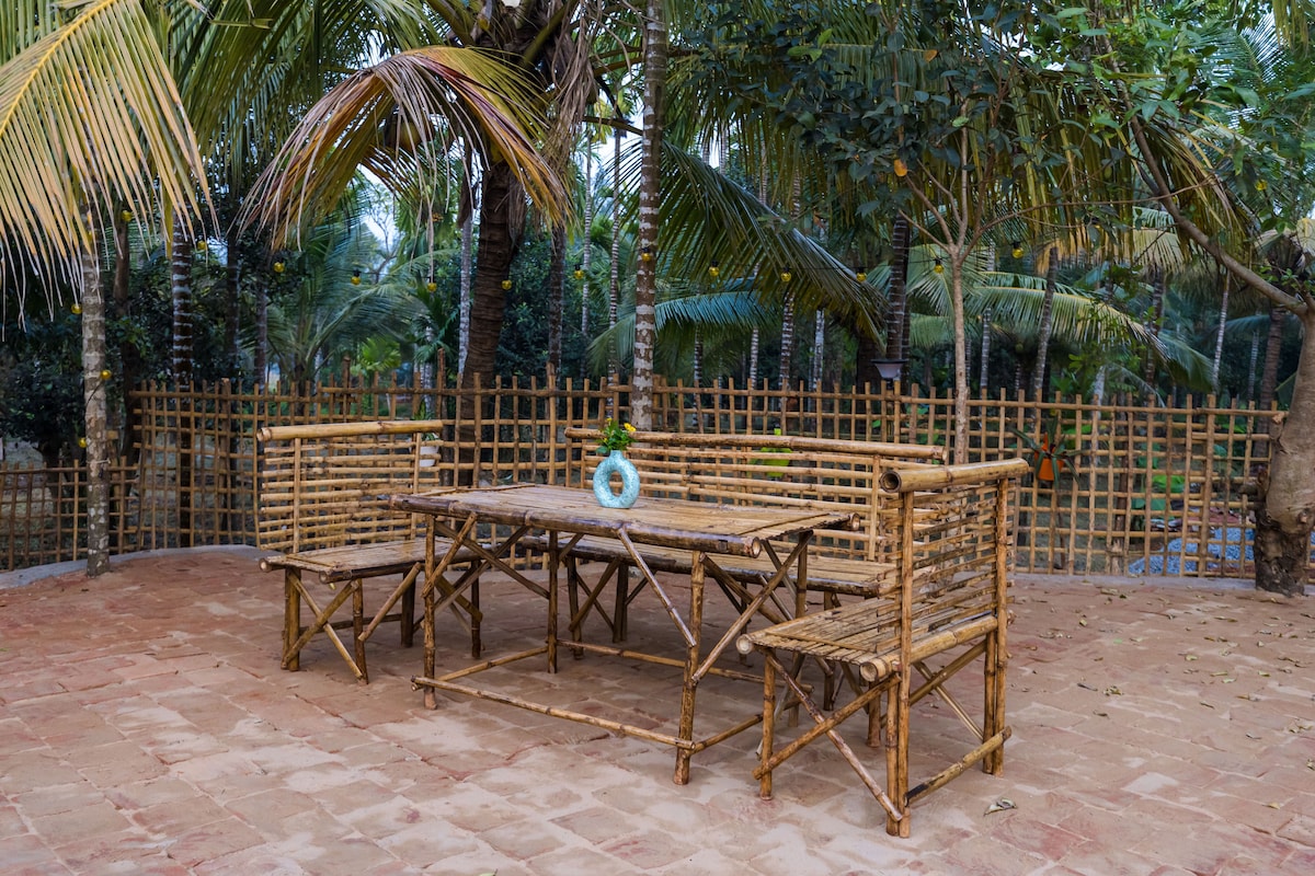 A bamboo dining set is positioned on a tiled patio, surrounded by lush greenery and palm trees. The table is flanked by benches, creating a welcoming outdoor dining space. A decorative vase is placed in the center of the table, adding a touch of charm.