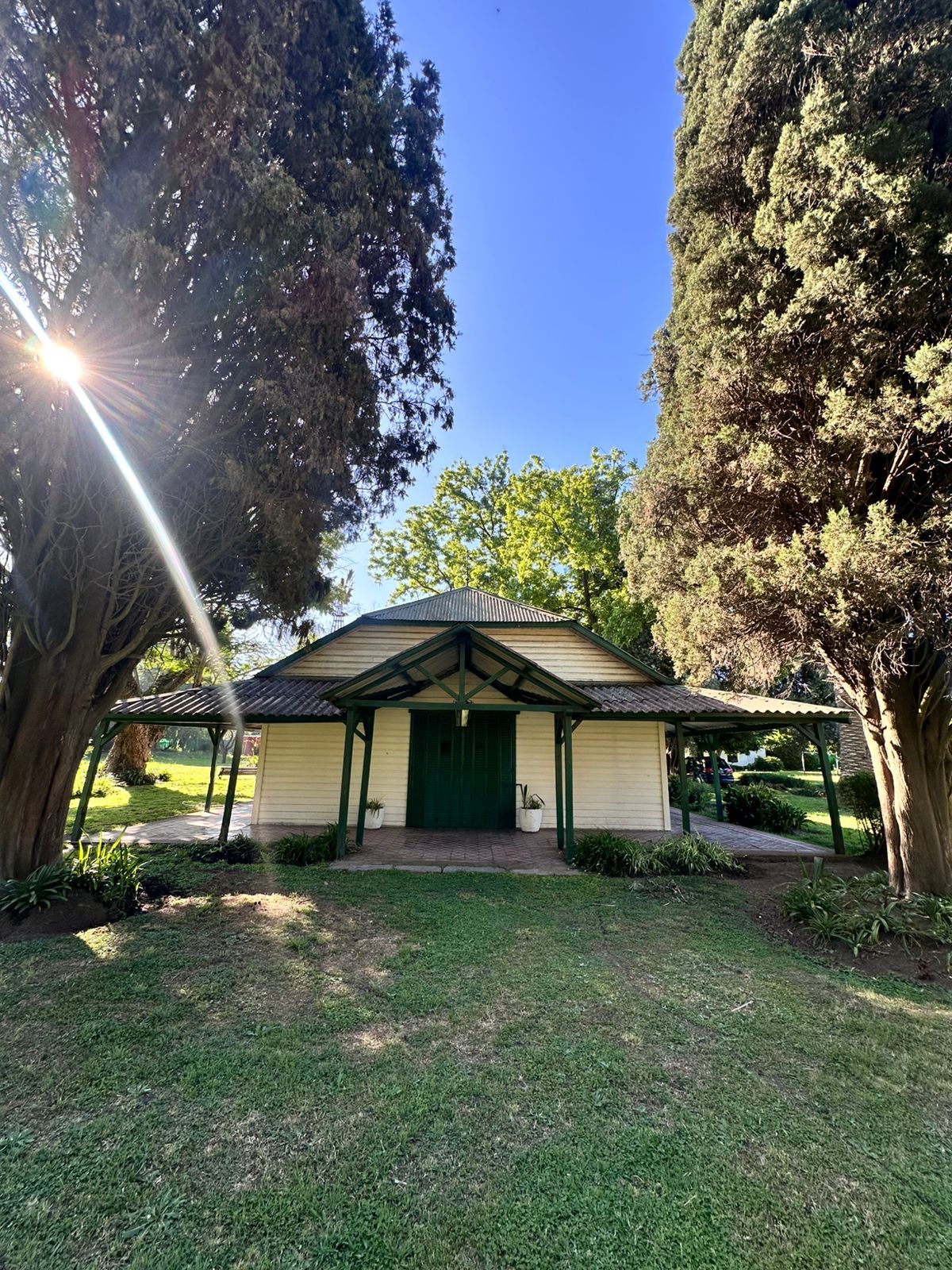 A charming structure is framed by tall trees, showcasing a green and white exterior. Sunlight filters through the branches, creating dappled patterns on the grass. The entrance features double doors, inviting guests into the tranquil environment of the surrounding park.